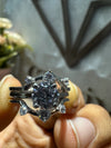 Silver ring with a large central gemstone held between fingers against a blurred background